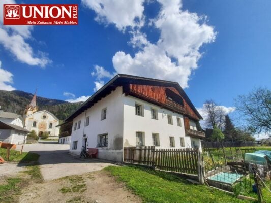 Großes Haus im ländlichen Stil mit weißen Wänden und dunklem Holzdach unter blauem Himmel mit Wolken, umgeben von Natur und Garten, Blick auf Berge im Hintergrund.
