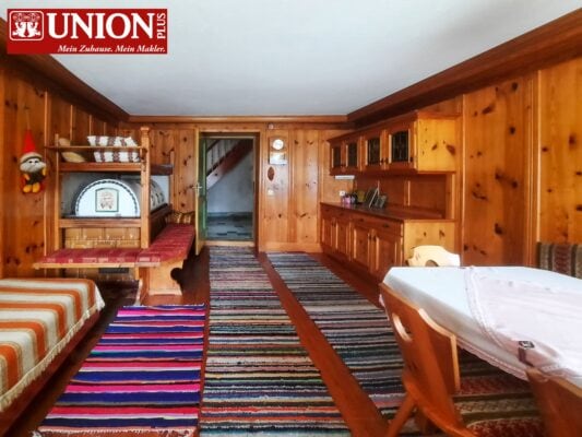 Gemütliches Wohnzimmer mit holzverkleideter Wand, bunten Teppichen, Ess- und Sitzbereich, perfekt für eine Familien- oder Gemeinschaftsunterkunft im Alpenstil.
