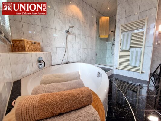 Sauberes Badezimmer mit Badewanne und Dusche, modern gestaltet mit weißen Fliesen und schwarzem Marmorfußboden, Handtücher auf Holzständer, Heizkörper und Rattankorb, ideale Wohlfühloase, in der Nähe der Union Plus Immobilienwebseite.
