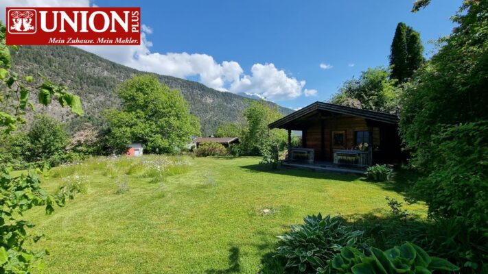 Schönes grünes Grundstück mit einer kleinen Hütte im Garten, umgeben von Bäumen und Bergen im Hintergrund. Perfekt für Natur- und Immobilienliebhaber, die Ruhe und idyllische Landschaft suchen.