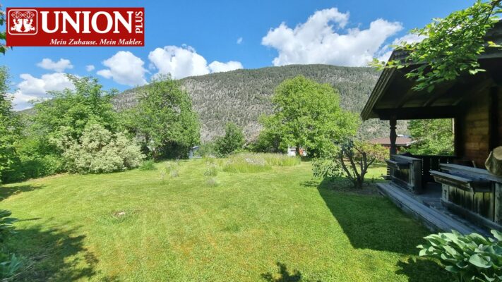 Garten in ländlicher Umgebung mit grünem Rasen, Bäumen und Berg im Hintergrund, sonniges Sommerwetter mit blauen Himmel und Wolken, Holzhaus im Vordergrund, ideal für Natur- und Immobilien-Suchmaschinen.