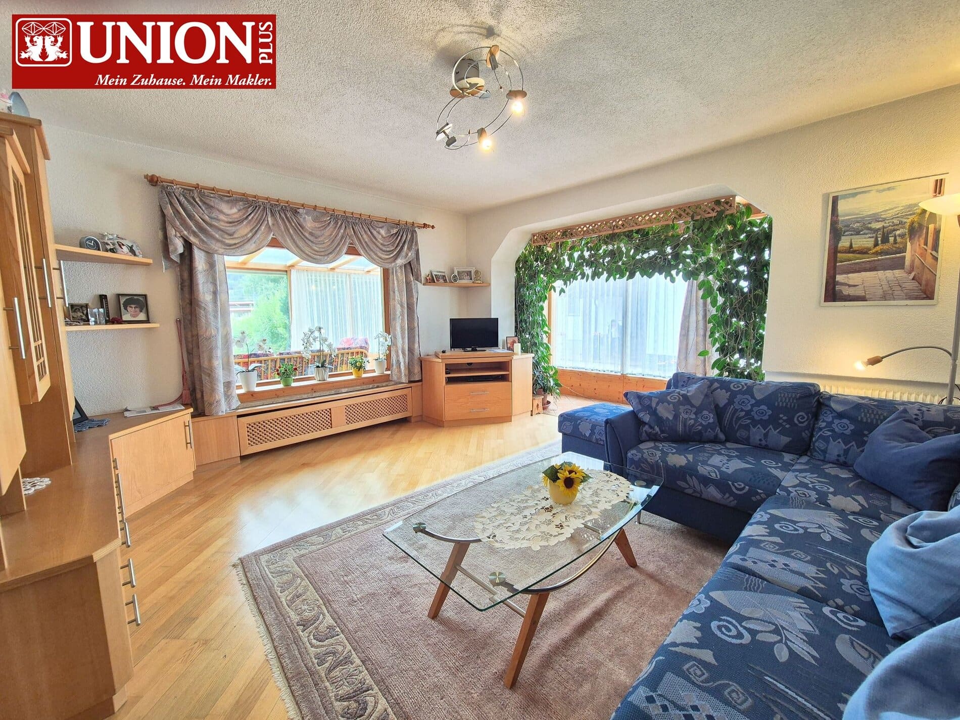 Gemütliches Wohnzimmer mit großen Fenstern, Holzmöbeln, blauer Couch, Pflanzen und natürlicher Beleuchtung für einladende Atmosphäre.