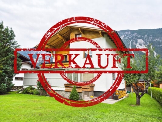Häuser im Tiroler Stil mit Grünfläche und Bergen im Hintergrund, verkauft mit "Verkauft" Stempel.