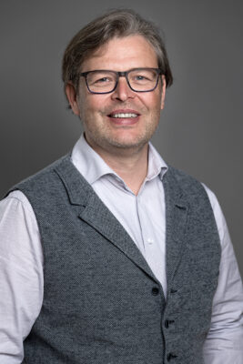 Freundlicher Mann mit Brille und grau melierter Frisur, trägt weißen Hemd und grauen Anzug, Corporate Portrait, Berufsbild, moderne Business-Fotografie.