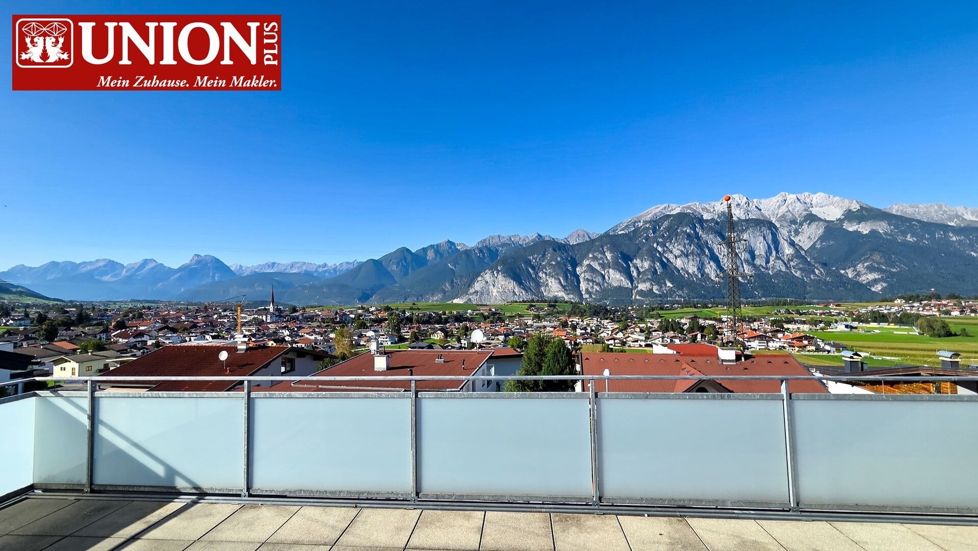 Hochwertiges Bild eines Bergpanoramas mit Blick auf eine österreichische Stadt und schneebedeckte Alpen im Hintergrund, aufgenommen von einem Balkon aus. Ideal für Inhalte zu Immobilien, Alpen, Berge, Stadtansichten und Natur in Österreich.