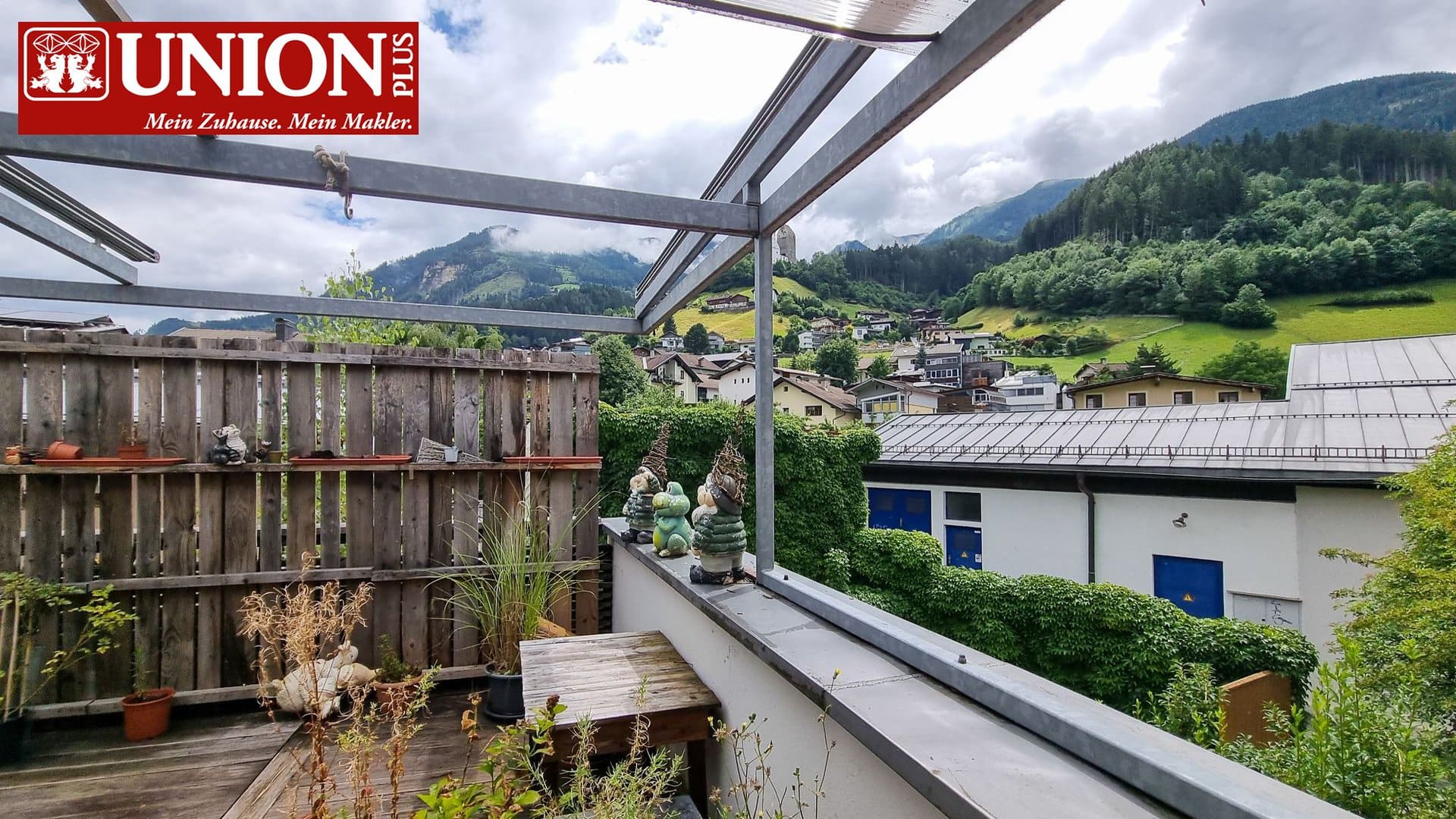 Gartenbalkon mit Blick auf die Alpen, umgeben von grünen Hügeln und Häusern, mit Dekorationsartikeln und Pflanzen, bietet eine idyllische Aussicht auf die umliegende Natur.