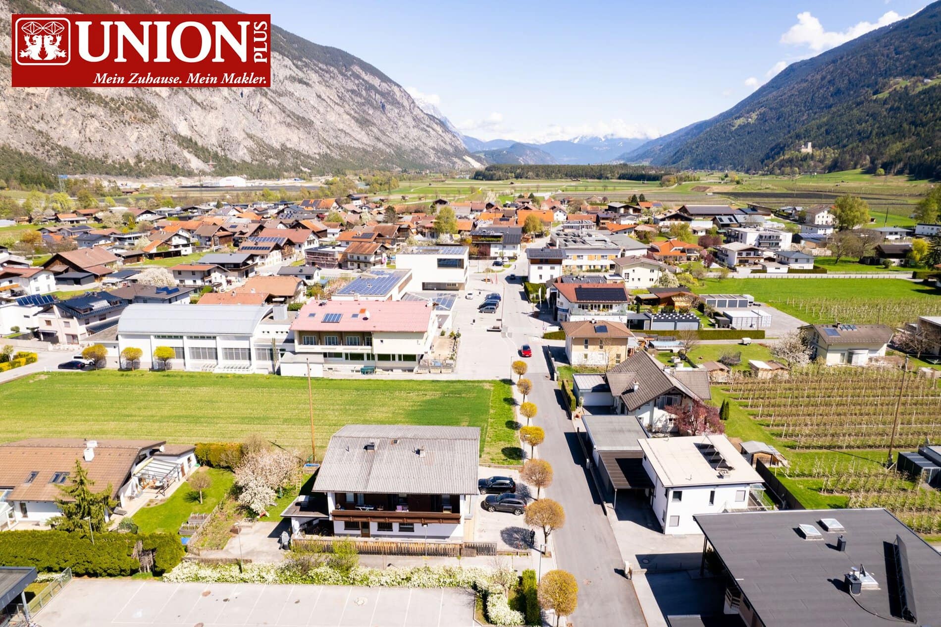 Luftaufnahme eines ruhigen Dorfes in den Bergen mit modernen Häusern, grünen Feldern und einer malerischen Bergkulisse im Hintergrund. Perfekt für Immobilien und Lebensqualität in ländlicher Umgebung.