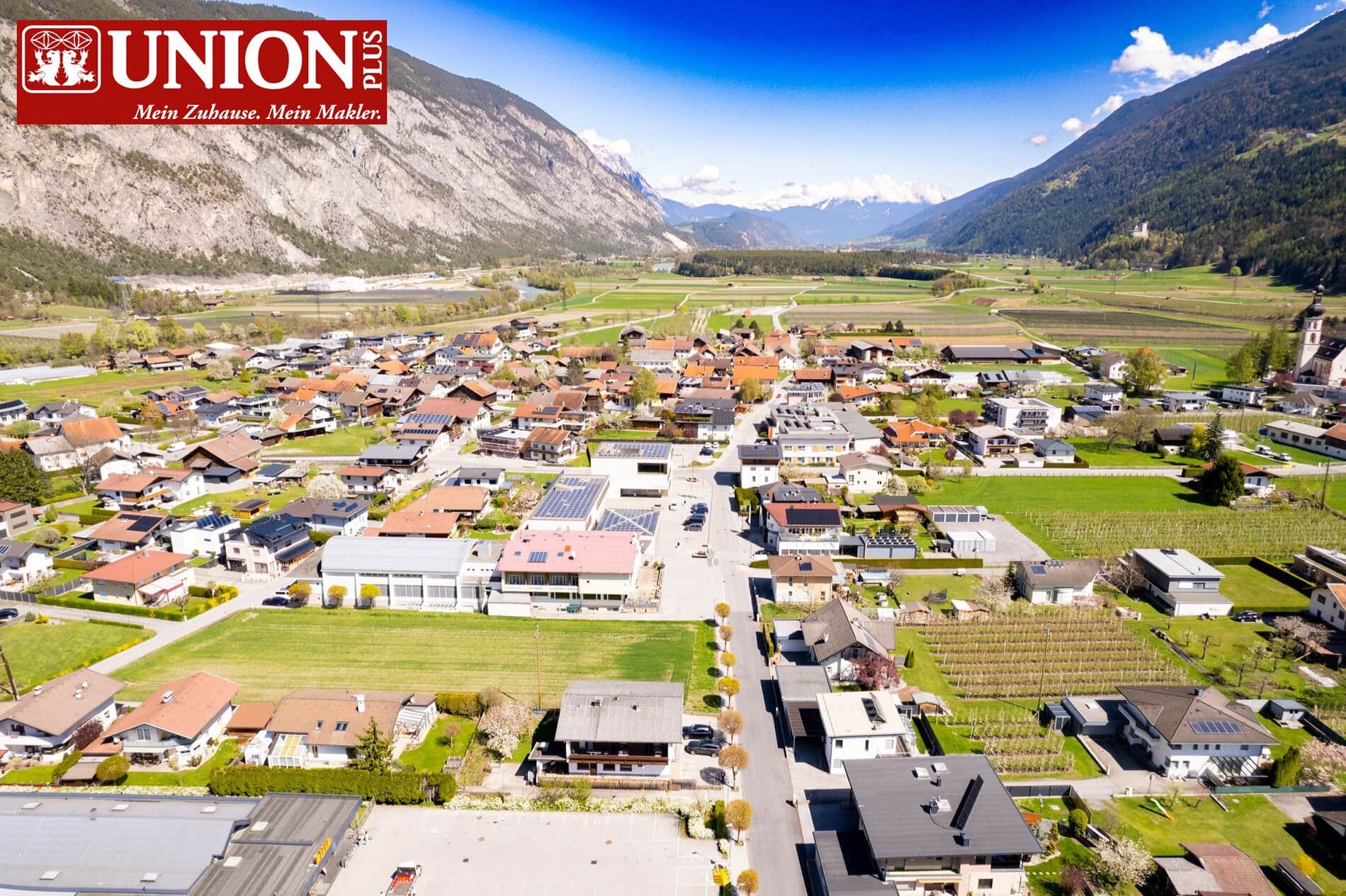 Luftaufnahme eines malerischen Dorfes im Alpenvorland mit Bergen im Hintergrund, grünen Feldern und modernen Häusern, symbolisiert Gemeinschaft und Lebensqualität.